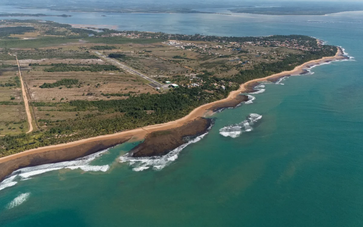 Vista aérea de uma costa com praia de areia, floresta densa, estrada reta, estruturas residenciais espalhadas e águas turquesas com ondas