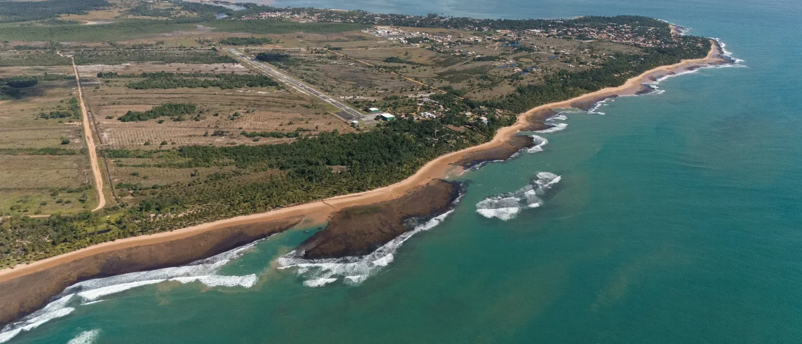 Vista aérea de uma costa com praia de areia, floresta densa, estrada reta, estruturas residenciais espalhadas e águas turquesas com ondas