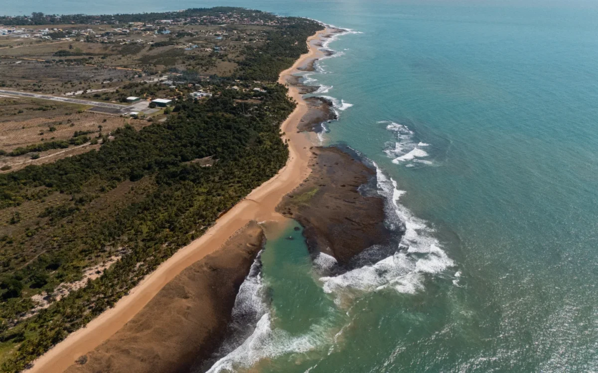 Vista aérea de costa com praia de areia, formações rochosas, vegetação densa, estrada, ondas e água azul-turquesa