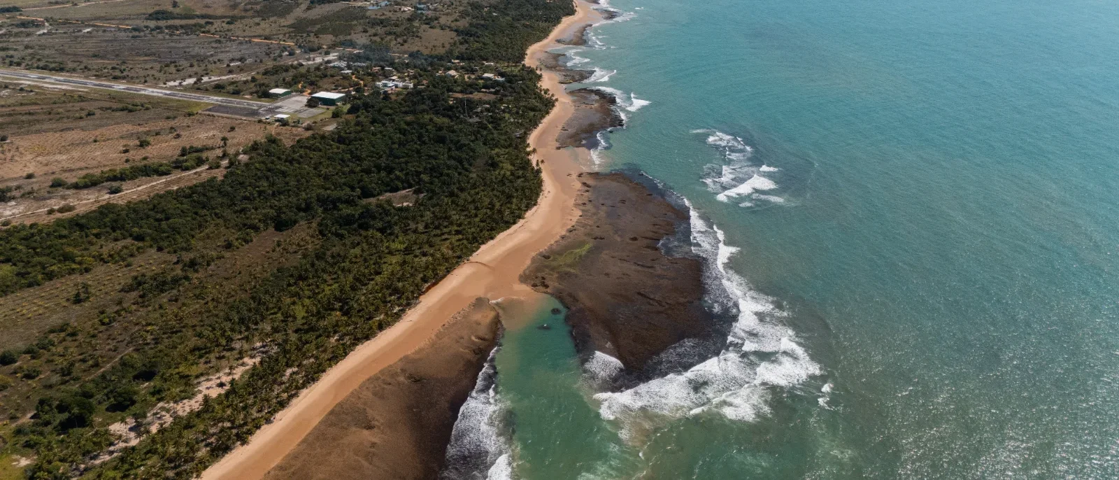 Vista aérea de costa com praia de areia, formações rochosas, vegetação densa, estrada, ondas e água azul-turquesa