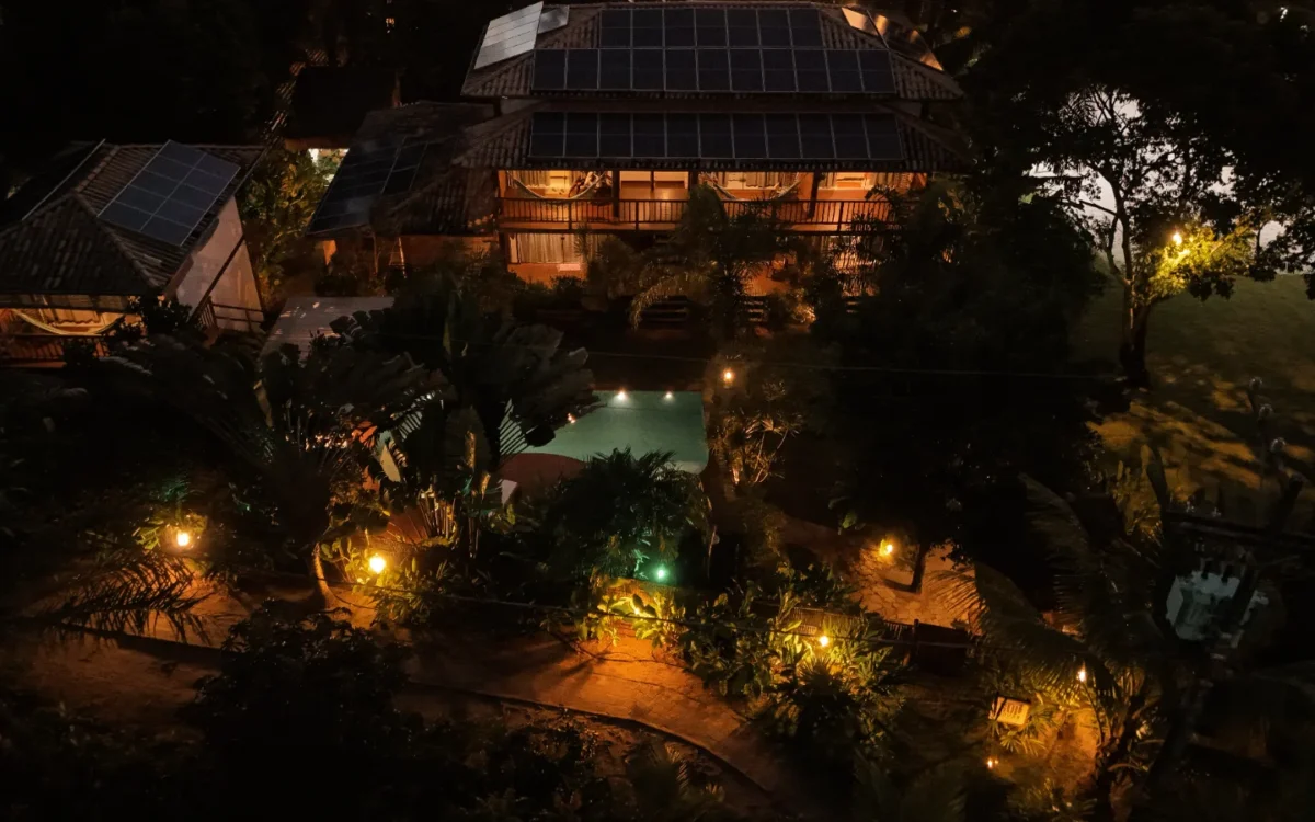 Vista aérea noturna de uma casa com piscina iluminada, painéis solares, jardim tropical e iluminação ambiente ao redor da propriedade