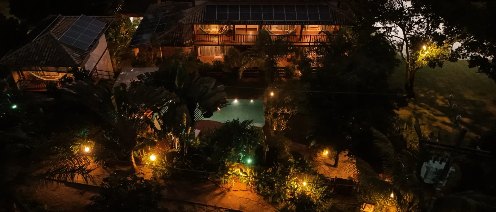 Vista aérea noturna de uma casa com piscina iluminada, painéis solares, jardim tropical e iluminação ambiente ao redor da propriedade