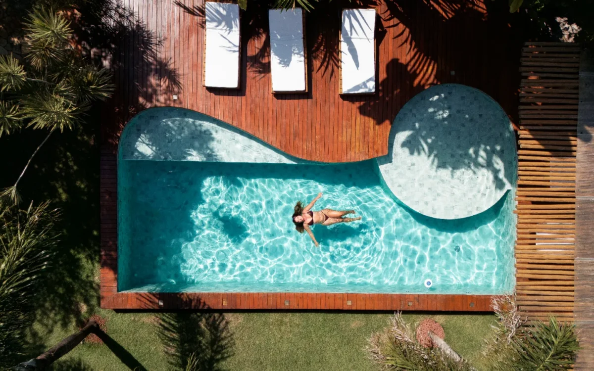 Pessoa flutuando em piscina em formato de onda com água cristalina, cercada por deck de madeira, janelas e plantas tropicais