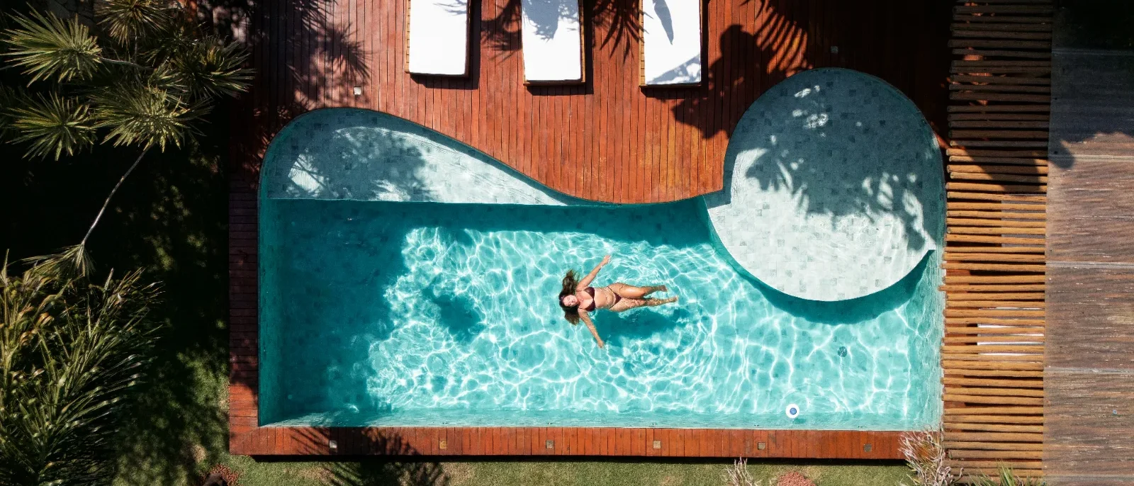 Pessoa flutuando em piscina em formato de onda com água cristalina, cercada por deck de madeira, janelas e plantas tropicais