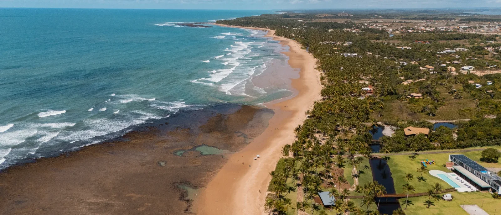 Vista aérea de uma praia com areia dourada, ondas, vegetação tropical, casarões e piscina à beira-mar sob céu azul