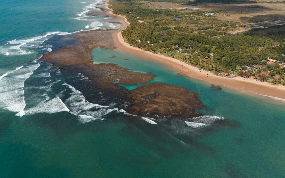 Vista aérea de praia tropical com areia dourada, lagoa azul-turquesa, formações rochosas escuras e floresta densa ao fundo