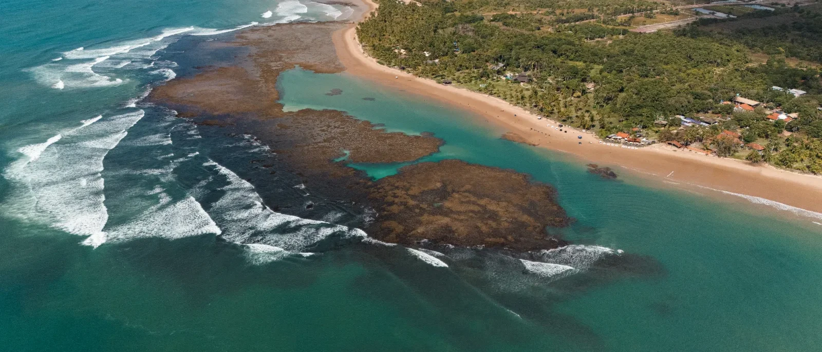 Vista aérea de praia tropical com areia dourada, lagoa azul-turquesa, formações rochosas escuras e floresta densa ao fundo