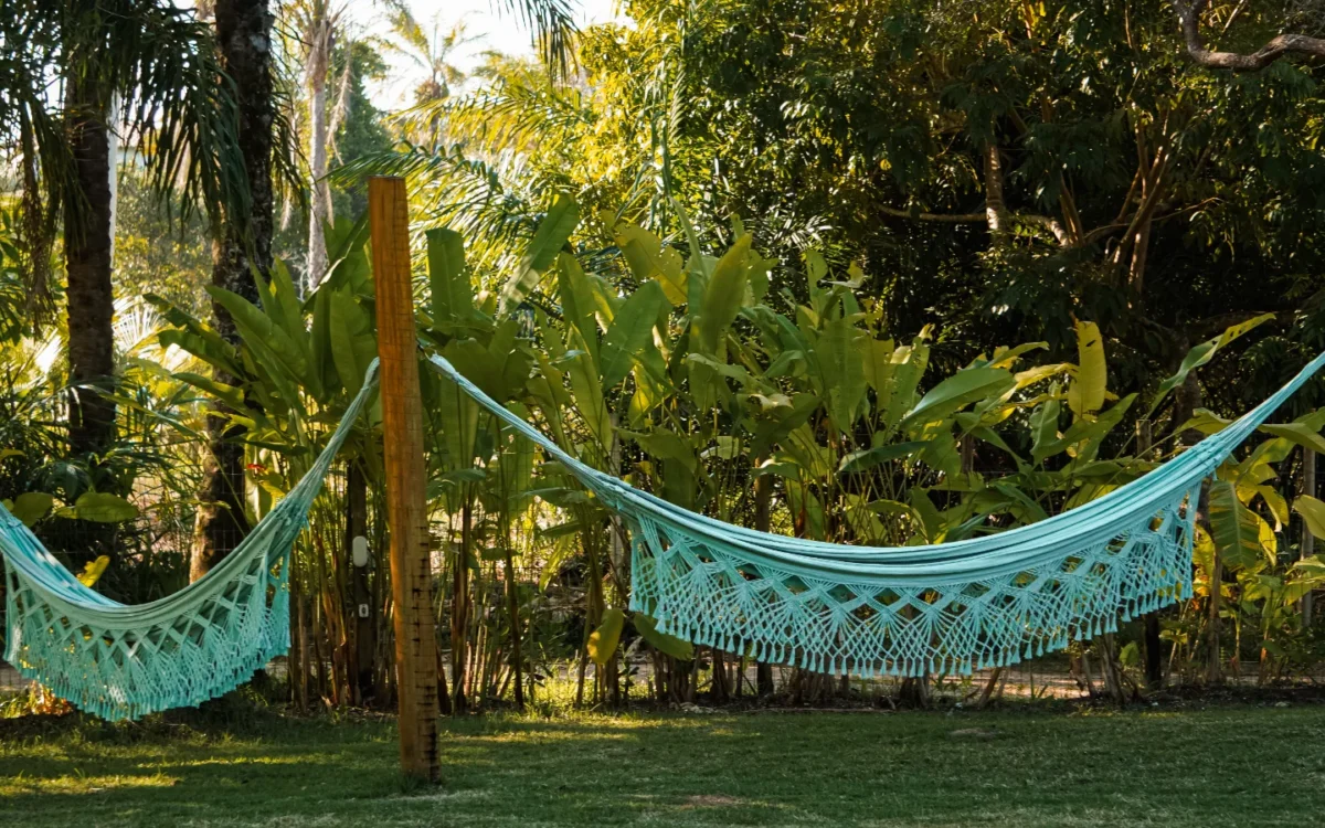 Duas redes de macramê azul turquesa penduradas entre árvores e postes em um gramado cercado por vegetação tropical