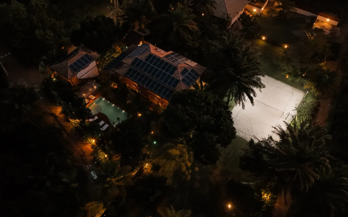 Vista aérea noturna de residência com painéis solares no telhado, piscina iluminada e área cercada por vegetação densa