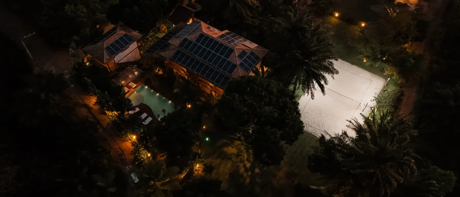 Vista aérea noturna de residência com painéis solares no telhado, piscina iluminada e área cercada por vegetação densa