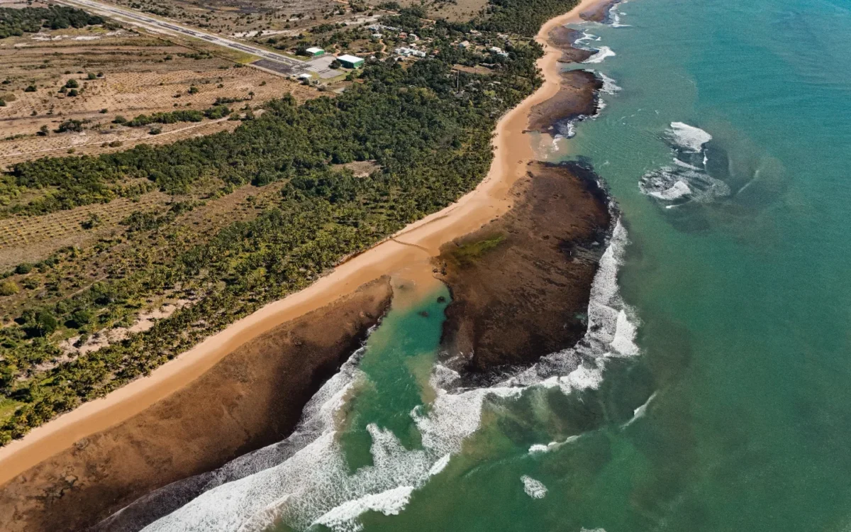 Vista aérea de costa com praia de areia dourada, formações rochosas, ondas do mar turquesa, vegetação verde e estrada na região superior
