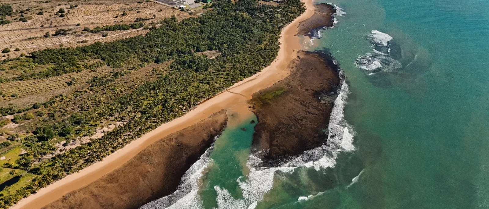 Vista aérea de costa com praia de areia dourada, formações rochosas, ondas do mar turquesa, vegetação verde e estrada na região superior