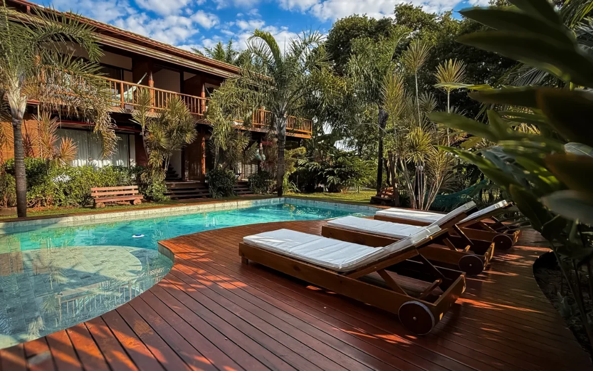 Casa de madeira com piscina azul cristalina, espreguiçadeiras brancas no deck de madeira, cercada por palmeiras e vegetação tropical sob céu azul
