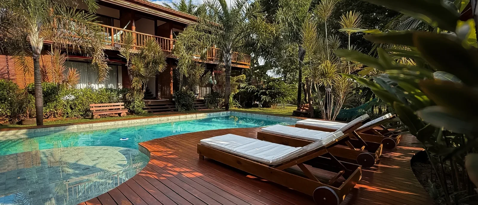 Casa de madeira com piscina azul cristalina, espreguiçadeiras brancas no deck de madeira, cercada por palmeiras e vegetação tropical sob céu azul
