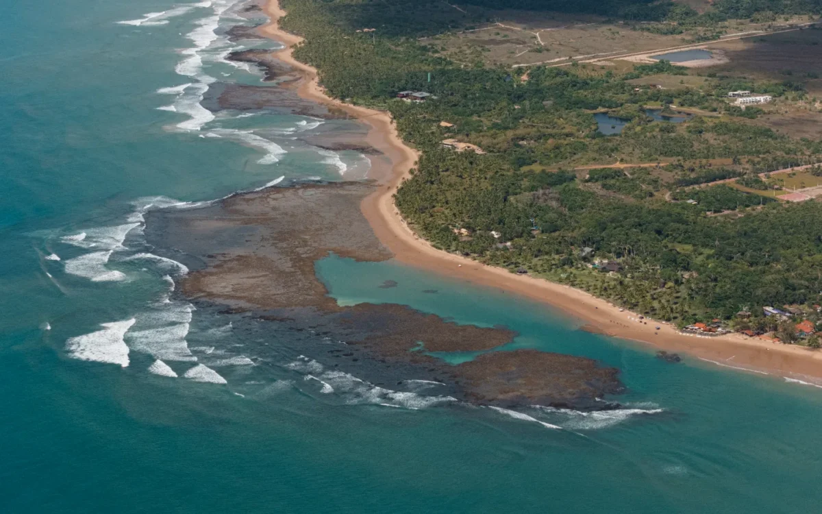 Vista aérea de uma praia com areia clara, lagoa turquesa, rochas vulcânicas escuras, ondas brancas e floresta tropical densa ao fundo