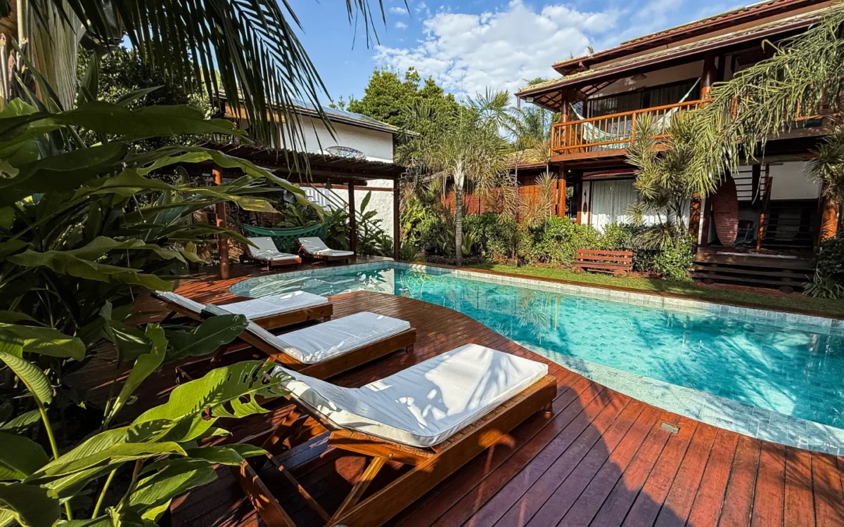 Resort tropical com piscina azul, espreguiçadeiras de madeira com toalhas brancas, vegetação exuberante e casarões de madeira ao fundo sob céu azul