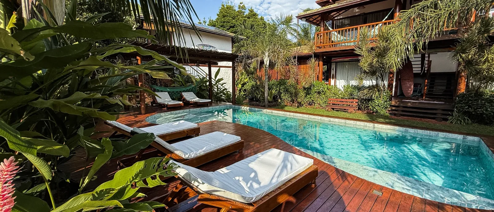 Resort tropical com piscina azul, espreguiçadeiras de madeira com toalhas brancas, vegetação exuberante e casarões de madeira ao fundo sob céu azul