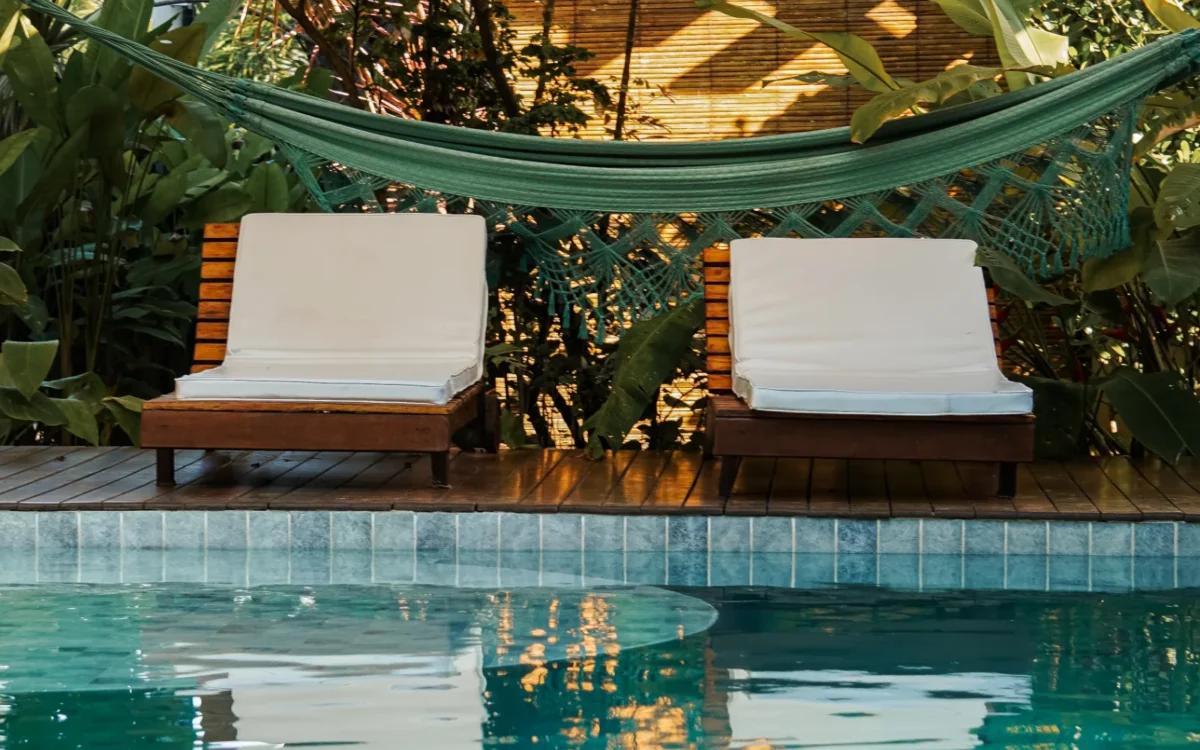 Piscina com água verde e azulejos, duas espreguiçadeiras com almofadas brancas, pergolado de madeira com rede verde e plantas ao redor