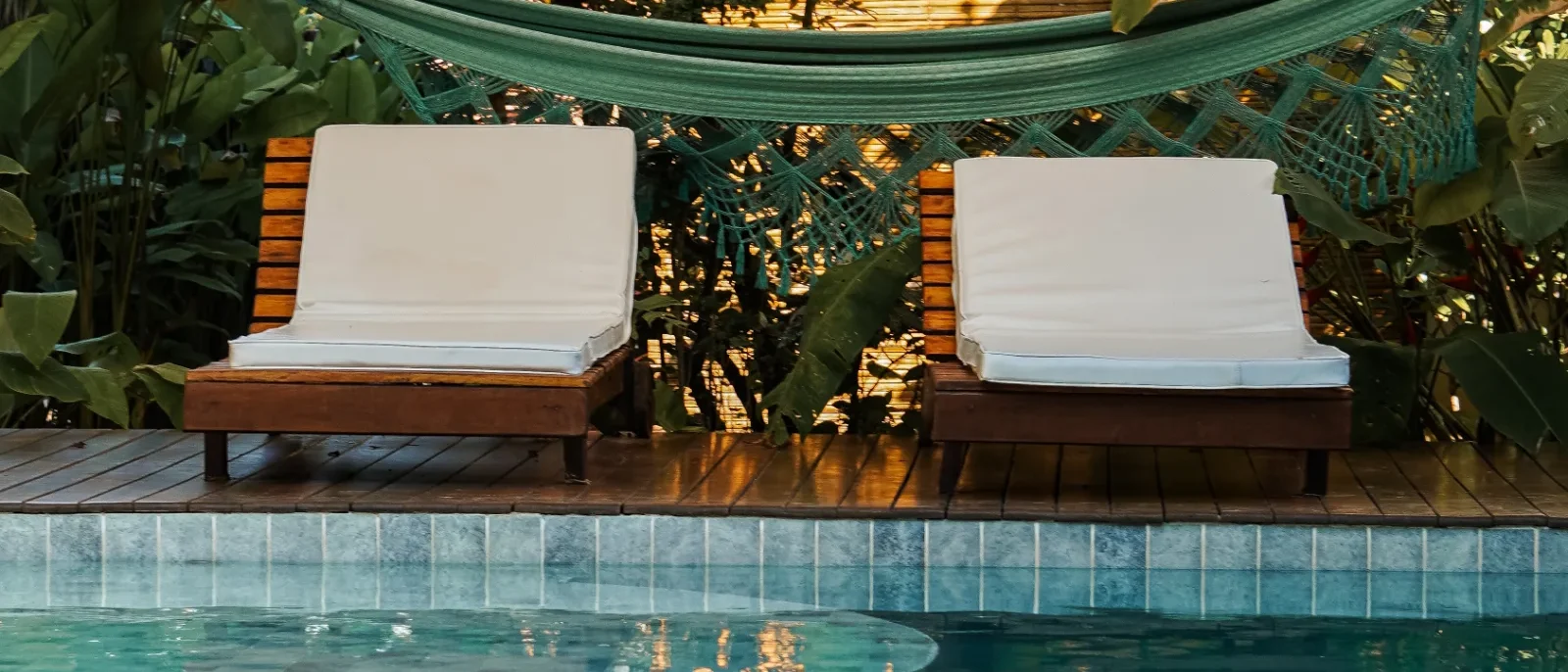 Piscina com água verde e azulejos, duas espreguiçadeiras com almofadas brancas, pergolado de madeira com rede verde e plantas ao redor