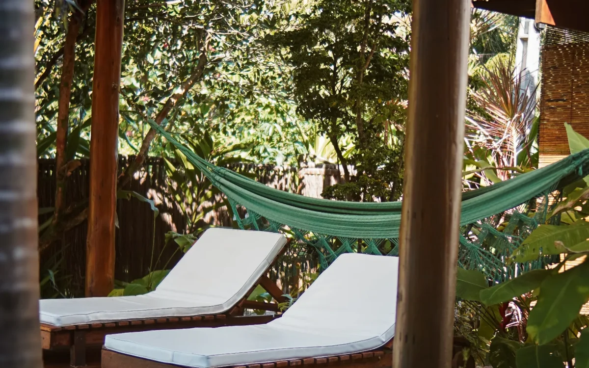 Varanda coberta com dois espreguiçadeiras brancas, piscina ao fundo, rede verde, plantas e vegetação tropical ao redor
