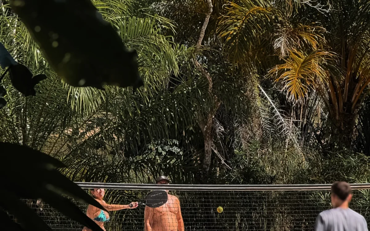 Três pessoas jogam vôlei de praia em uma quadra de areia com rede, cercadas por vegetação tropical e palmeiras