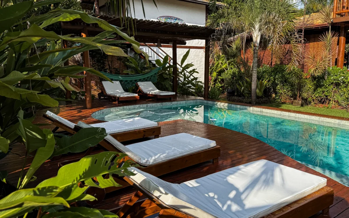 Piscina com água azul-turquesa cercada por espreguiçadeiras de madeira com colchões brancos, plantas tropicais, árvores de palma e construções de madeira ao fun