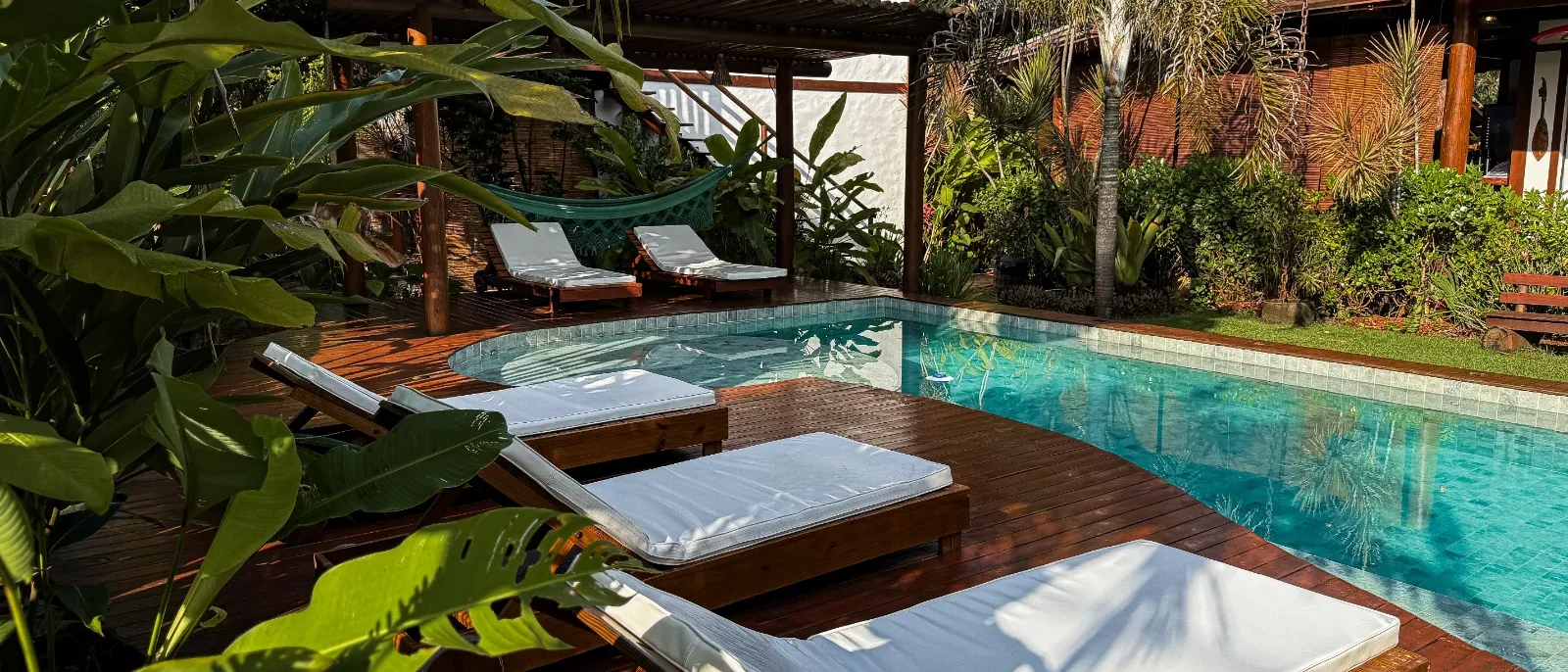 Piscina com água azul-turquesa cercada por espreguiçadeiras de madeira com colchões brancos, plantas tropicais, árvores de palma e construções de madeira ao fun