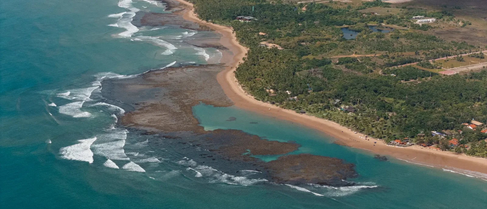 Vista aérea de uma praia com areia clara, lagoa turquesa, rochas vulcânicas escuras, ondas brancas e floresta tropical densa ao fundo