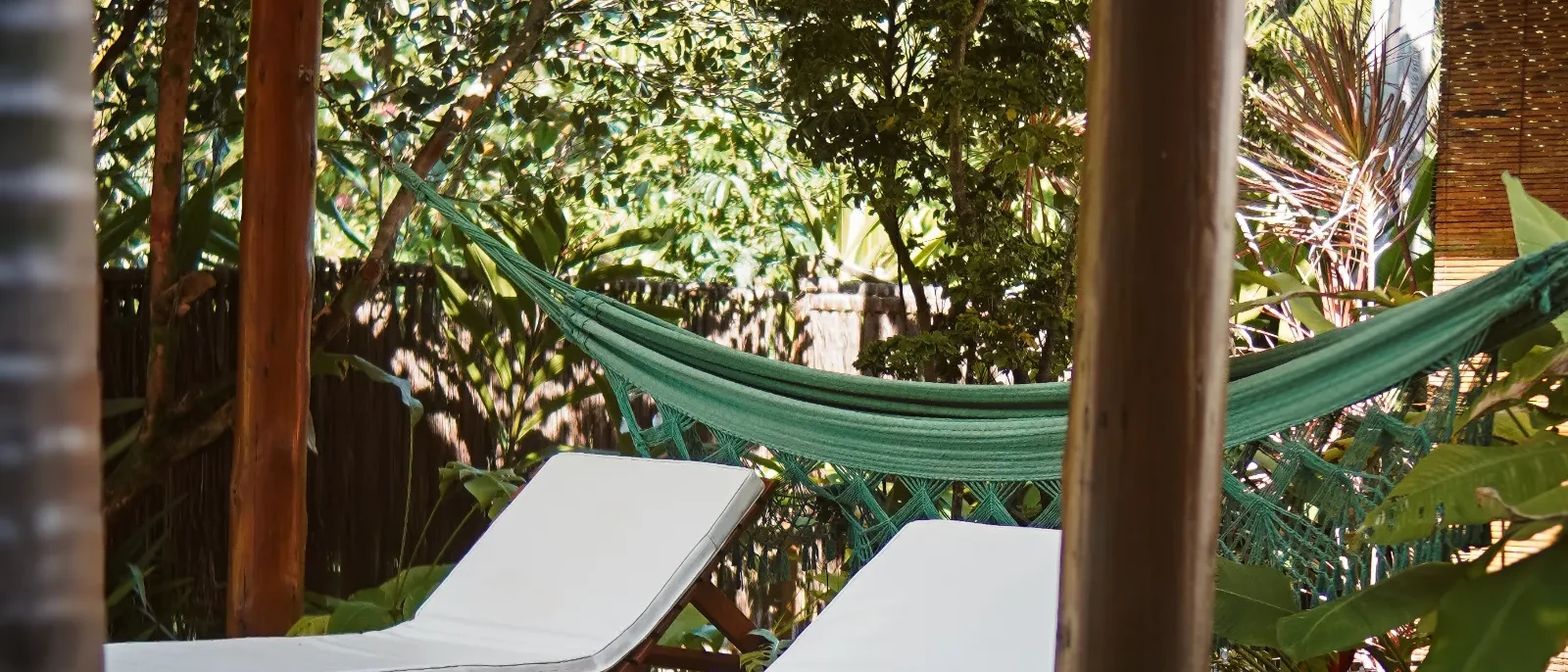 Varanda coberta com dois espreguiçadeiras brancas, piscina ao fundo, rede verde, plantas e vegetação tropical ao redor