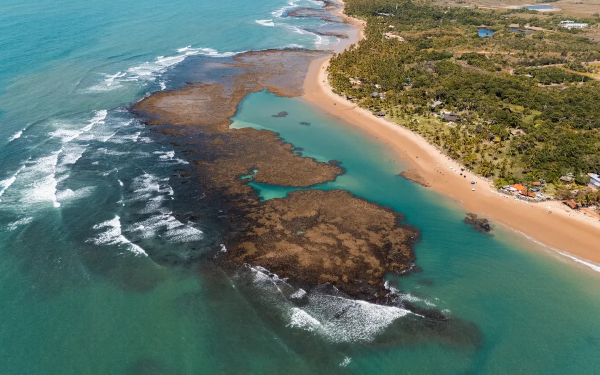 Vista aérea de praia com areia dourada, lagoa de água turquesa, rochas e vegetação tropical ao fundo, com ondas do oceano ao lado
