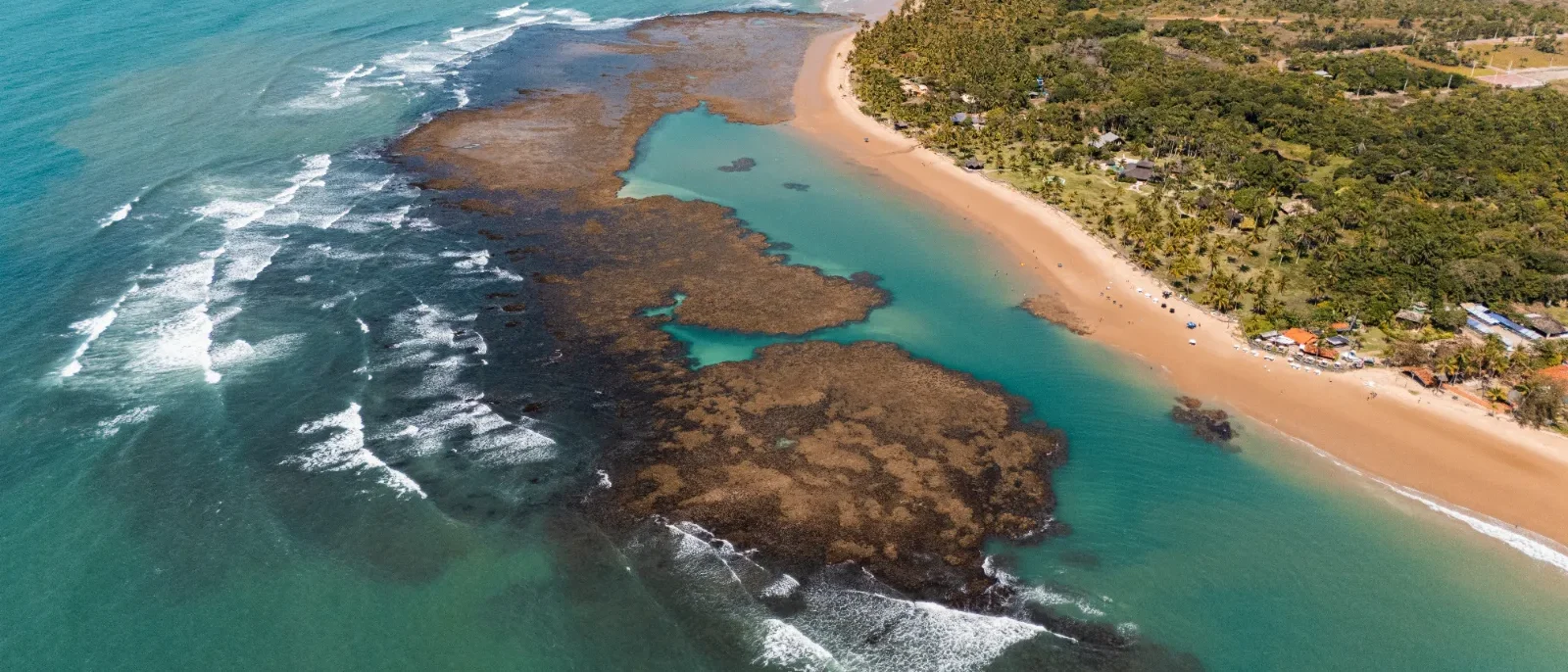 Vista aérea de praia com areia dourada, lagoa de água turquesa, rochas e vegetação tropical ao fundo, com ondas do oceano ao lado