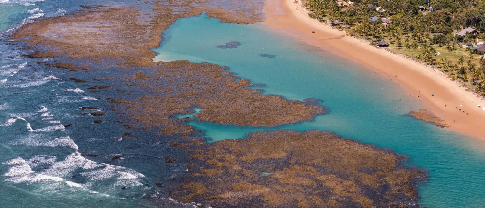 Vista aérea de praia com areia dourada, água turquesa, rochas marrom-avermelhadas e vegetação densa à direita, com ondas quebrando na costa