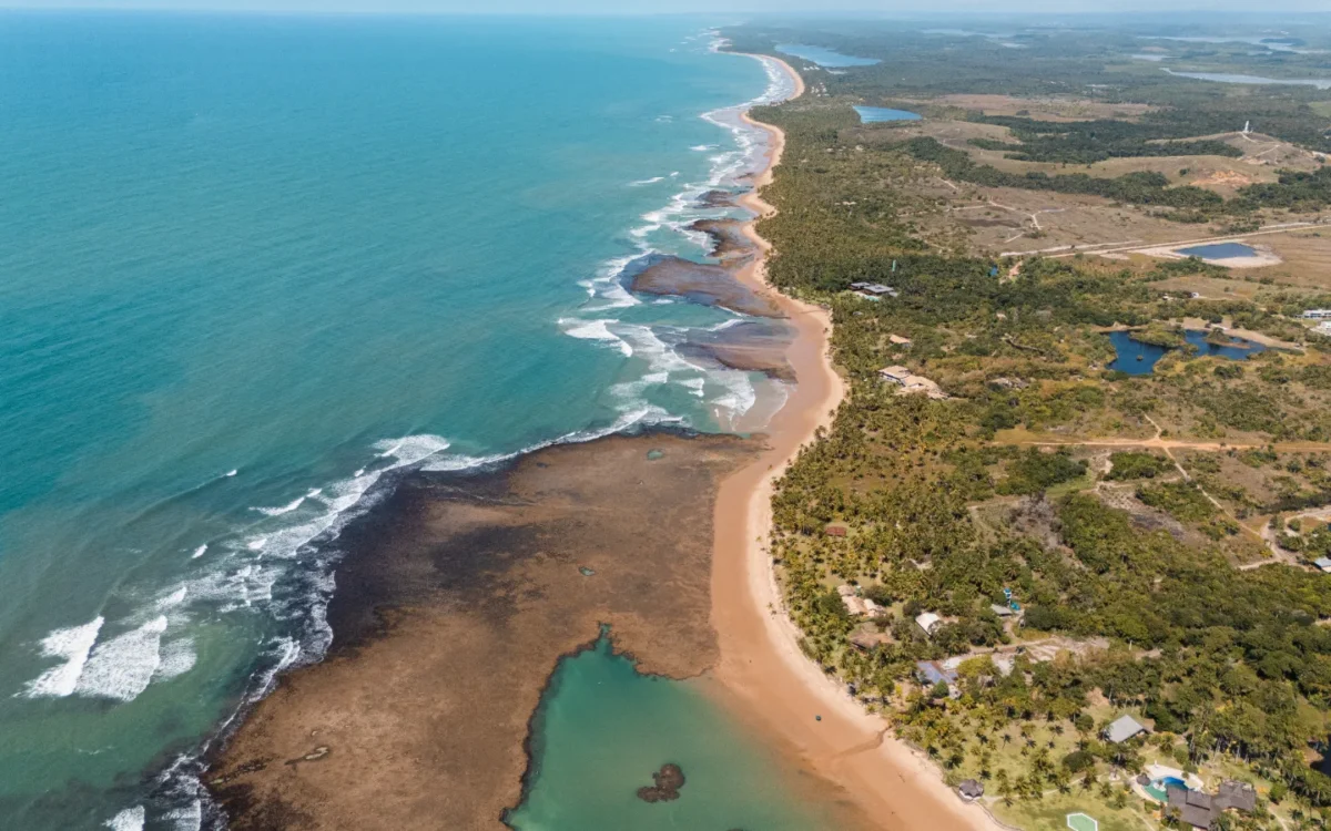 Vista aérea de uma costa com praia de areia clara, vegetação densa, lagoas azuis e ondas quebrando na água turquesa