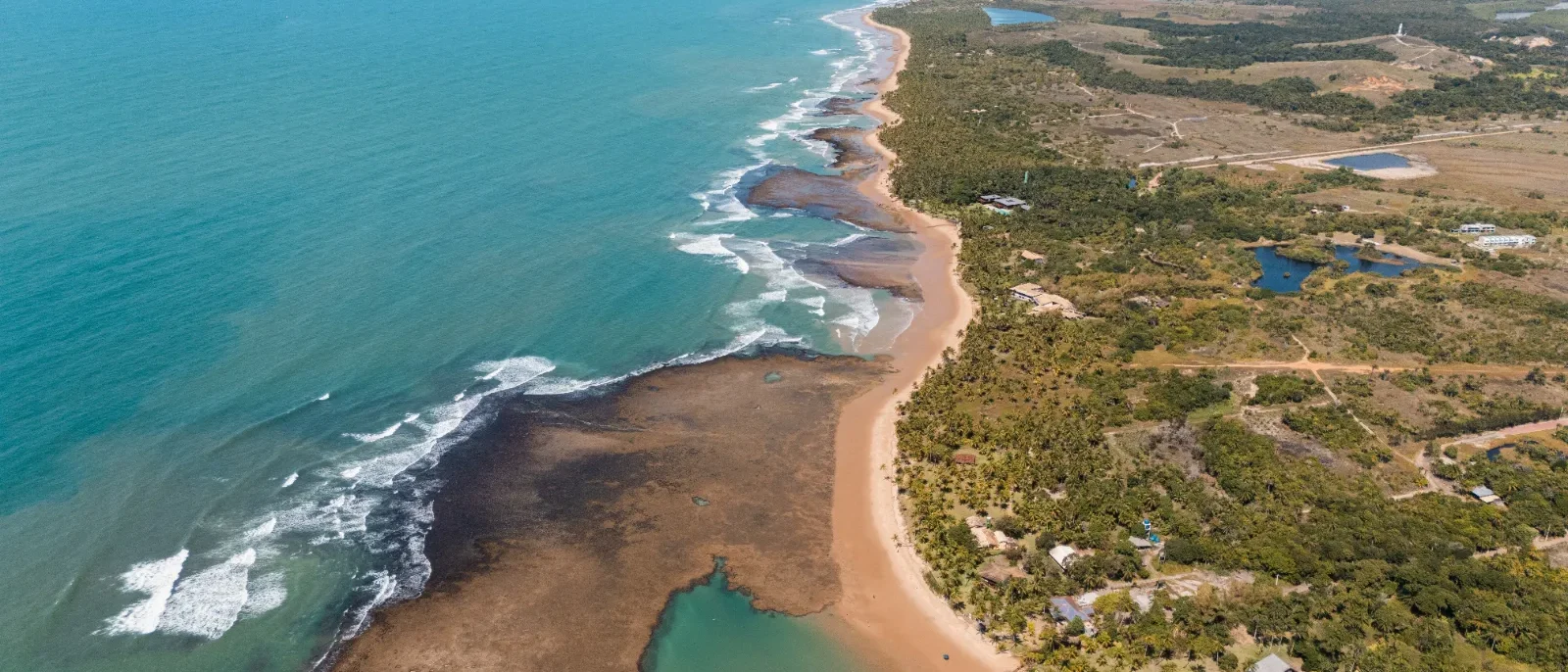 Vista aérea de uma costa com praia de areia clara, vegetação densa, lagoas azuis e ondas quebrando na água turquesa